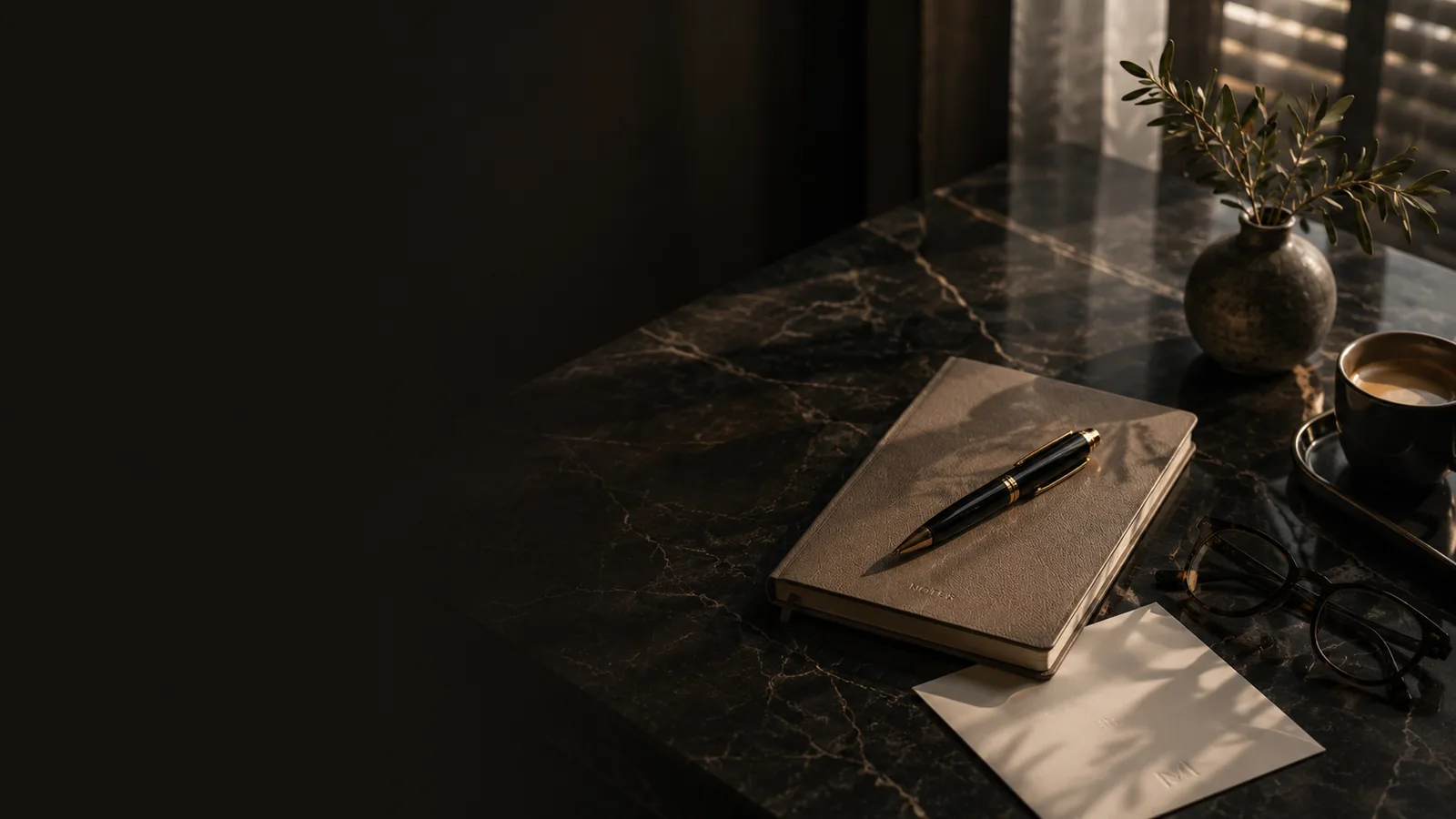 Bureau en marbre sombre avec carnet et stylo, branche d’olivier et café, lumière tamisée